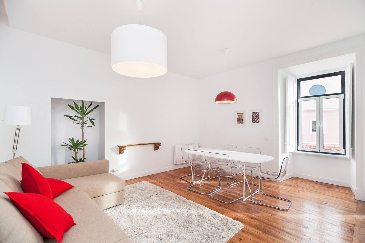 Una sala de estar moderna con un sofá beige, cojines rojos, una mesa de comedor blanca con sillas de metal, una alfombra blanca y un gran ventanal con vistas.