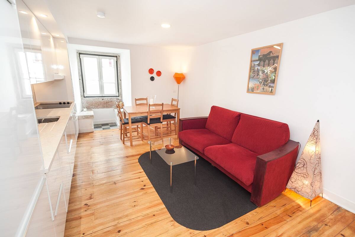 Una sala moderna con un sofá rojo, una mesa de comedor con sillas y una cocina al fondo.