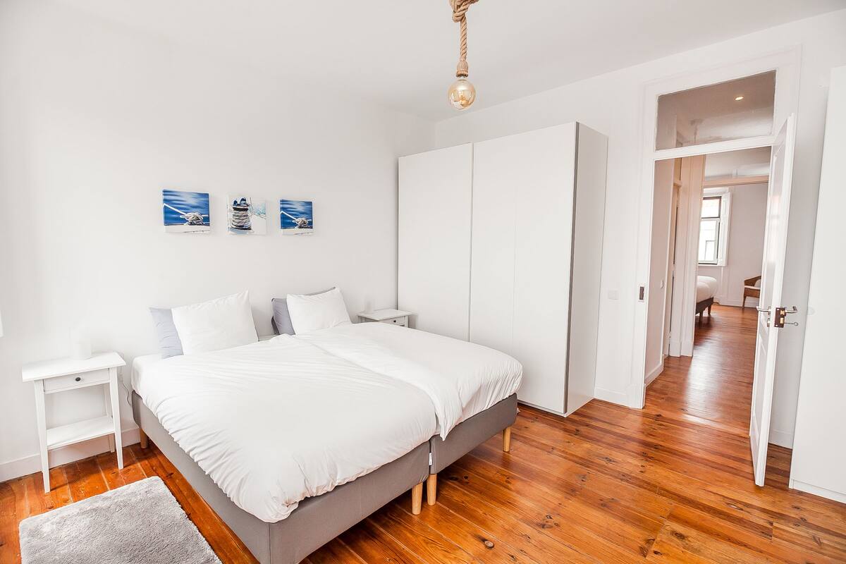 Un dormitorio con una cama grande, paredes blancas, suelo de madera y un pasillo que conduce a otra habitación.