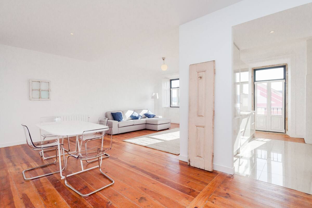 Una habitación amplia con piso de madera, una mesa de comedor blanca con sillas, un sofá con cojines y un gran ventanal que proporciona luz natural.