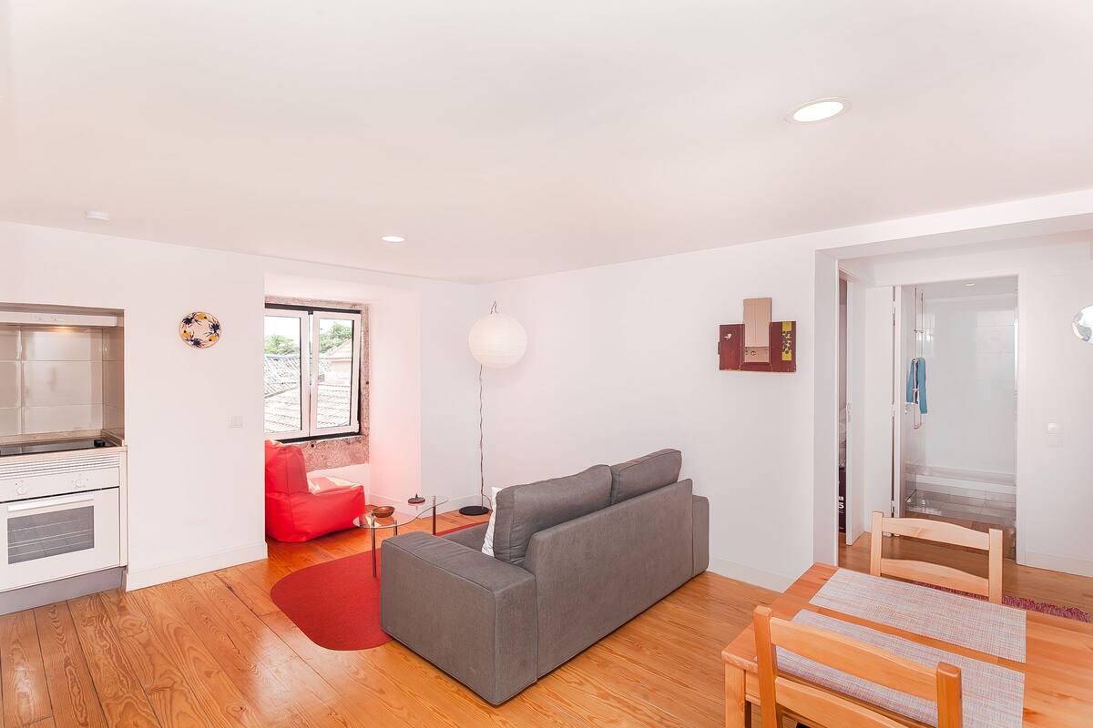 Una sala moderna con un sofá gris, una mesa de comedor de madera y una silla roja. Hay una zona de cocina con estufa y ventana.