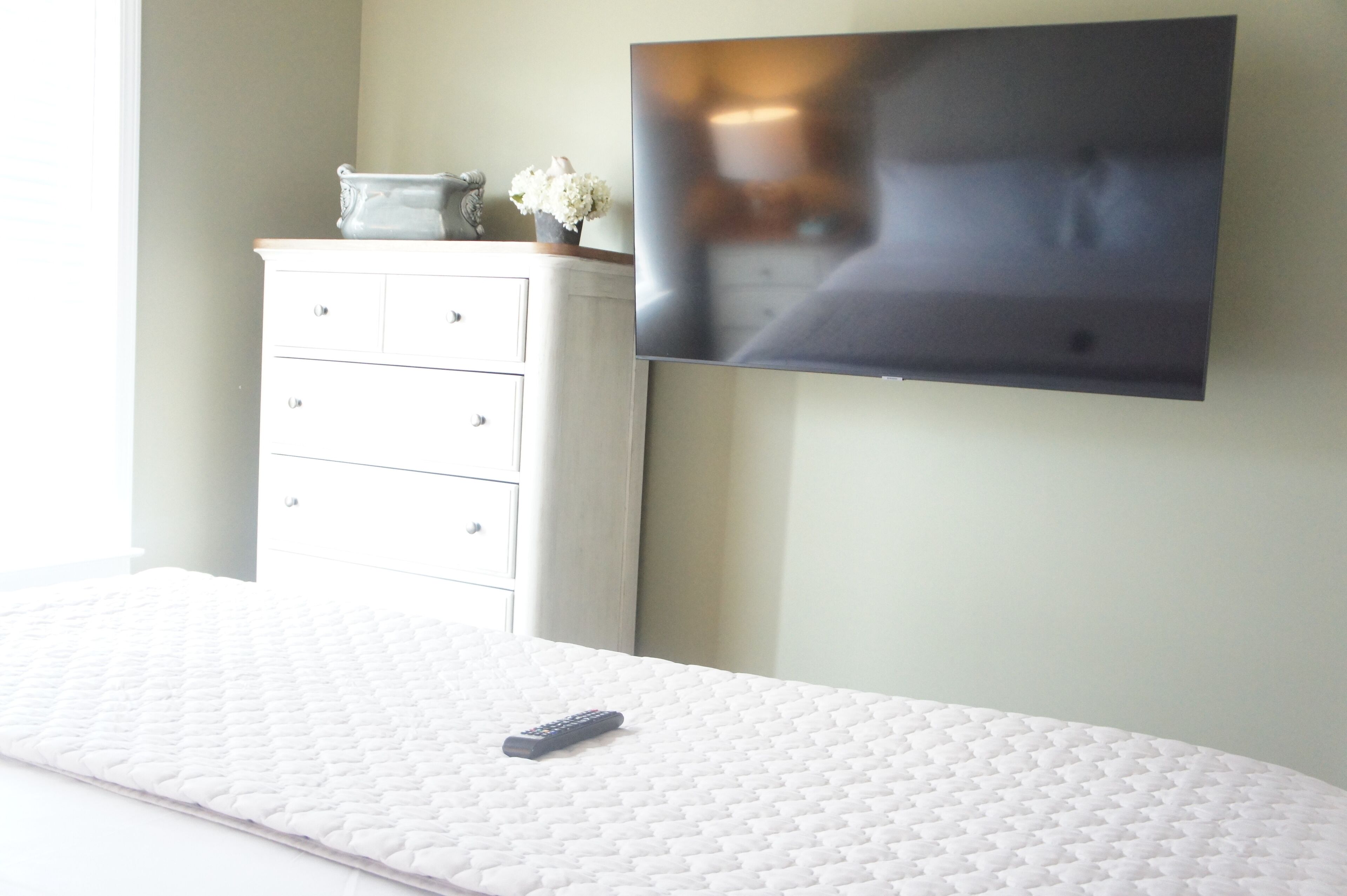 Un dormitorio con cama, televisor en la pared, cómoda y un jarrón con flores.