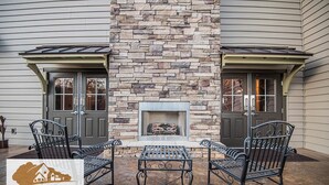 A stone fireplace with a fire, two metal chairs, and a table in front of a house with two doors. - Magnolia Creek Lodge (Cottonwood)