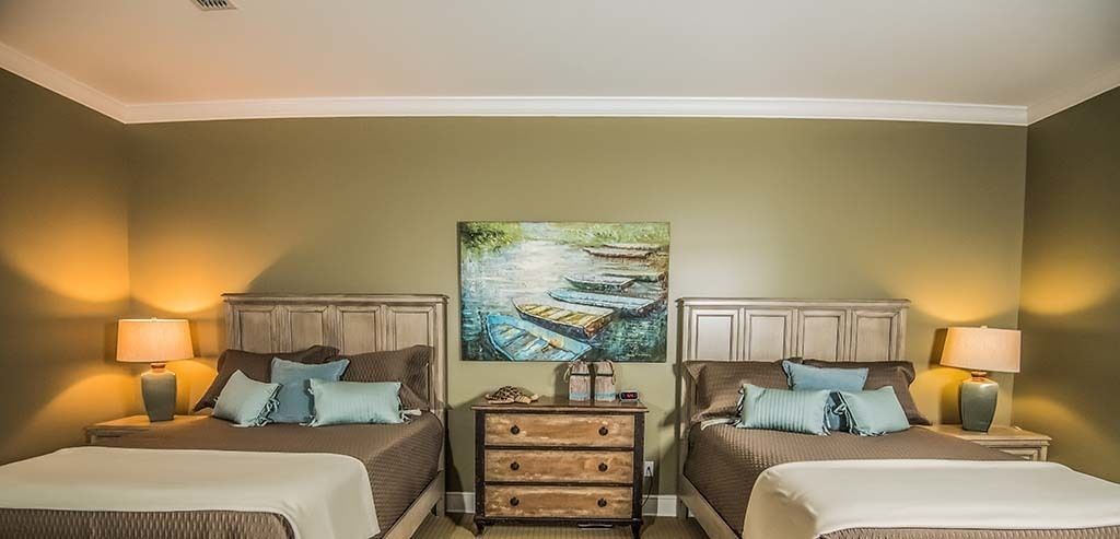 Habitación de hotel con dos camas, una mesita de noche con lámpara, un aparador y un cuadro de barcos en un río.