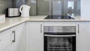 Moderni keittiö, jossa on ruostumattomasta teräksestä valmistetut kodinkoneet, kuten leivänpaahdin, vedenkeitin ja uuni. Keittiössä on valkoiset kaapit ja lasinen välitila.