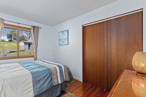 Un dormitorio con cama, una ventana con cortinas, una puerta de madera, un cuadro en la pared y una lámpara sobre una mesa.