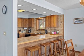 A kitchen with wooden cabinets, a large island with bar stools, a clock, and a sign on the wall. - Teak Village 1 - 54 - Three Bedroom House, Sleeps 8 (Lake Ozark)