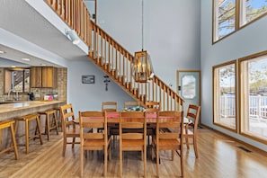 Un comedor con mesa y sillas de madera, una escalera con barandillas de madera y una cocina con pileta y alacenas.
