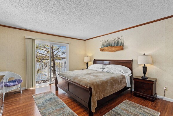 A bedroom with a large bed, two bedside tables, a chair, a balcony with a view, and a wooden headboard.