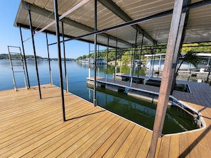 Een houten terras met metalen hekwerk met uitzicht op kalm water, liggende boten en een overdekt gebied met uitzicht op het water.