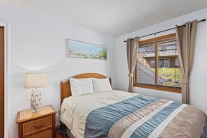 Un dormitorio con cama, mesita de noche, lámpara, ventana con cortinas y un cuadro en la pared.