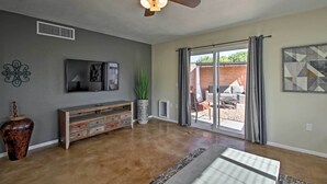 A spacious living room with a ceiling fan, a flat-screen TV, a wooden entertainment unit, a large plant, and a sliding glass door leading to an outdoor patio. - East Tucson Home w/ Backyard, Patio & Fire Pit (Tucson)