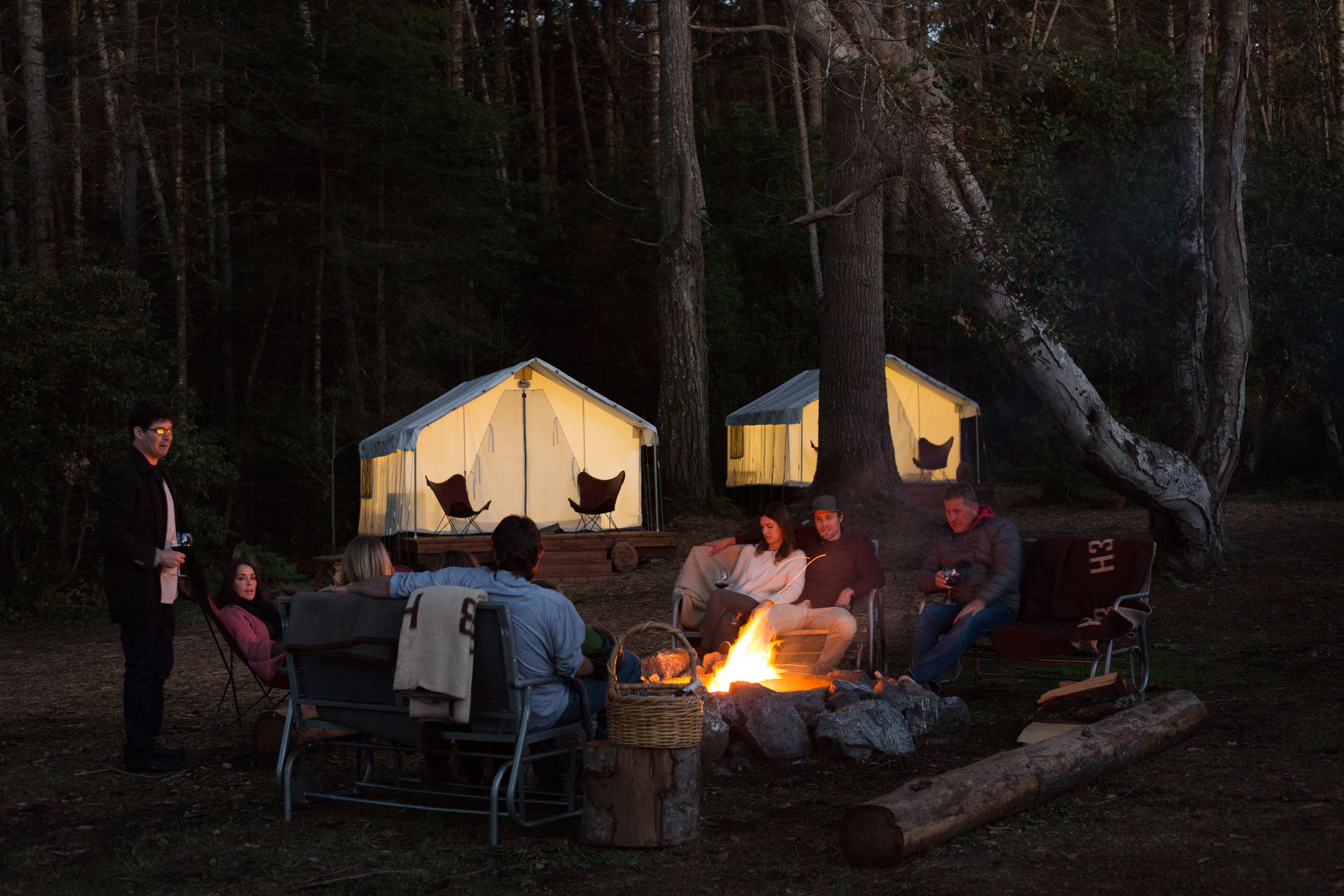 A group of people gathered around a campfire in a forested area with two small, tent-like structures in the background.