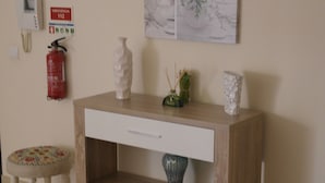 Un escritorio de madera con un cajón blanco, un estante pequeño y un florero con una planta. Hay un extintor y un cartel de salida de emergencia en la pared.