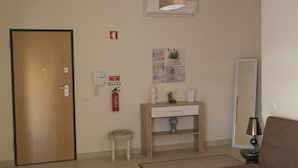Habitación de hotel con puerta de madera, un taburete blanco pequeño, una consola de madera con un jarrón, un extintor de incendios en la pared, un espejo y una lámpara.