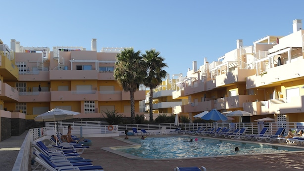Zona de piscina con sillas de descanso y sombrillas, rodeada de edificios de varios pisos con balcones.