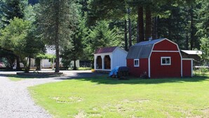 A red barn with a white door and a small attached shed, surrounded by trees and a gravel path. - Beautiful two bedroom one bath house overlooking the Siuslaw river. (Mapleton)