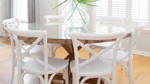 Een eethoek met een glazen tafel, omringd door witte stoelen, een potplant en een raam met jaloezieën.