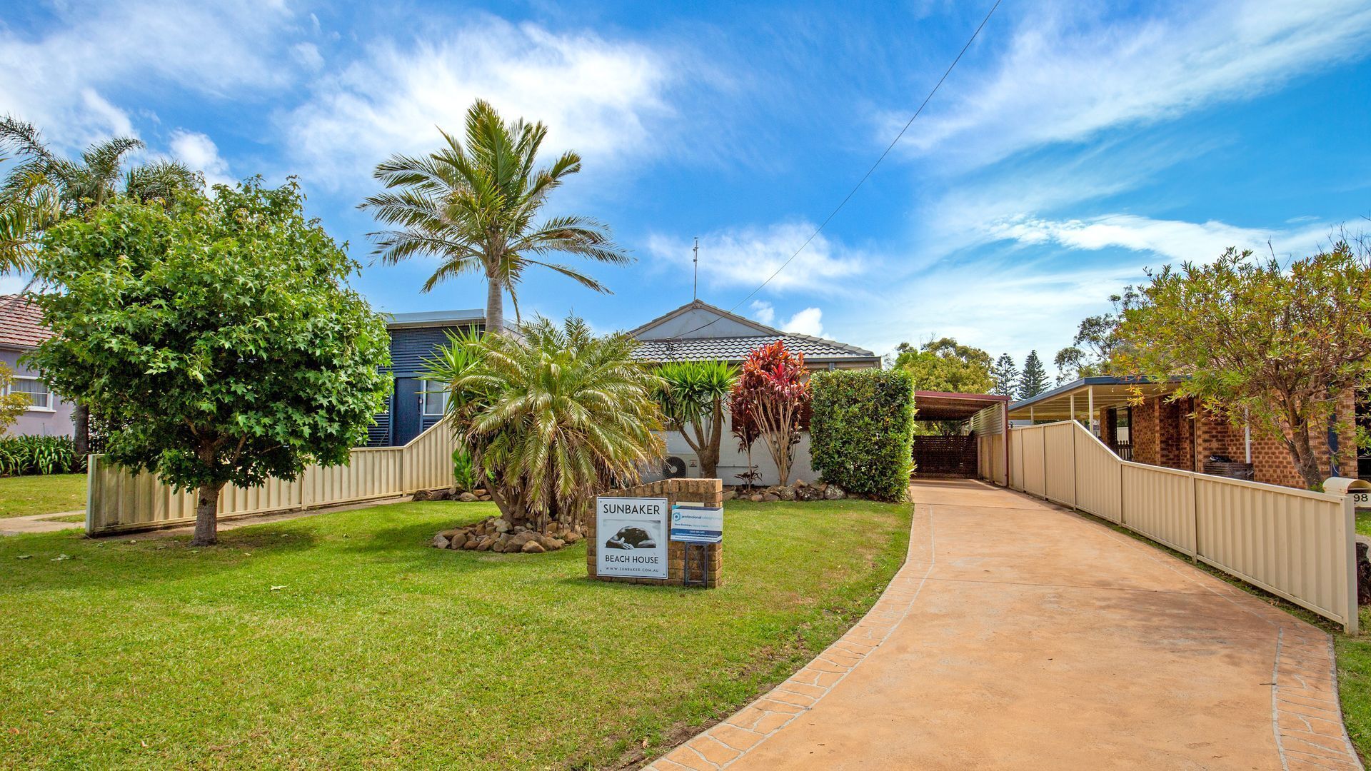 A residential property with a paved driveway, a sign indicating a beach house, and a variety of trees and shrubs.