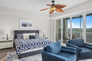 A bedroom with a bed, two nightstands, a ceiling fan, a large window, and a balcony with chairs. - Towers 9106 - Four Bedroom Apartment, Sleeps 10 (Osage Beach)