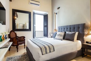 A bedroom with a large bed, a bedside table, a chair, a mirror, and a window with shutters.