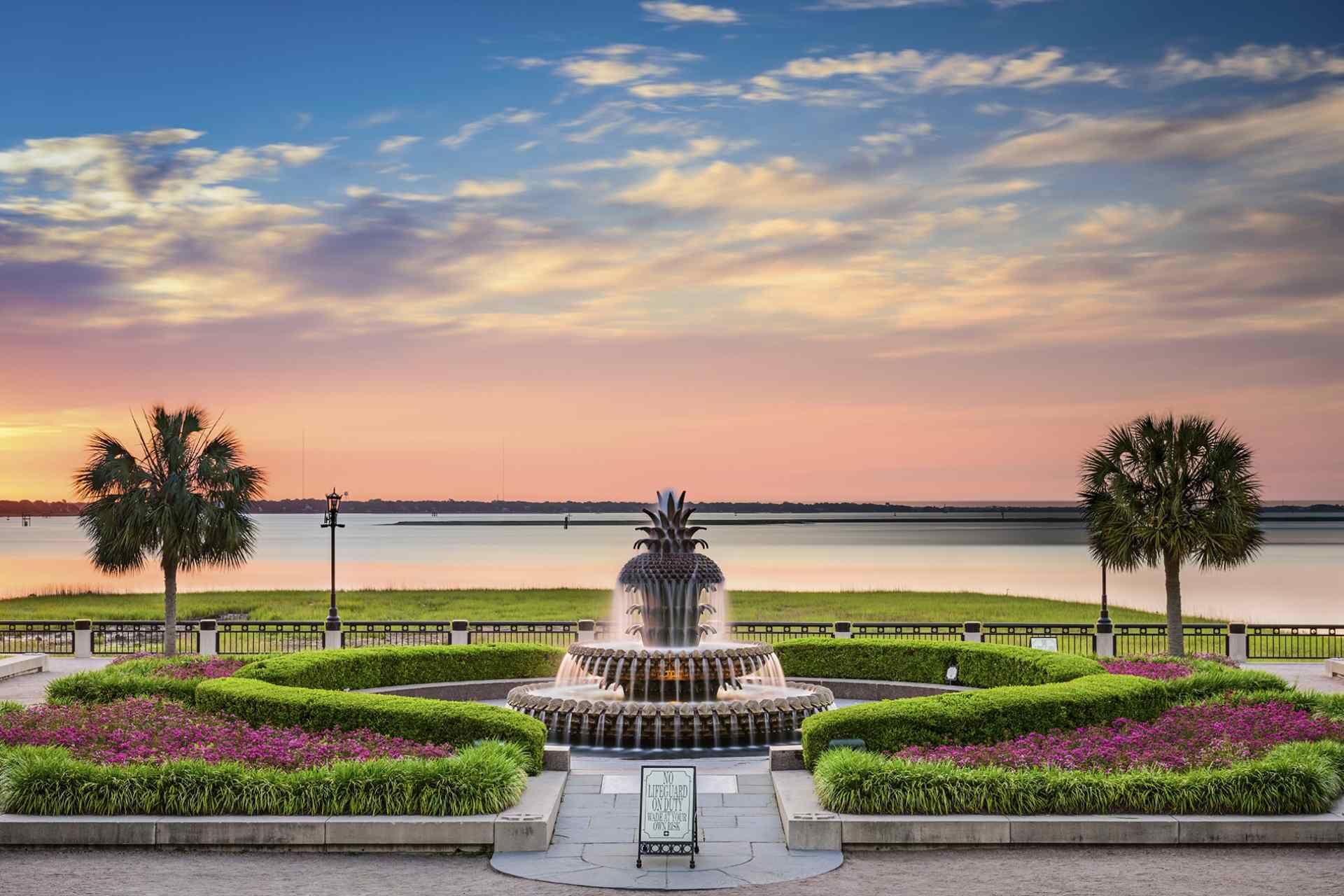 Una fuente rodeada de jardines cuidados y palmeras, con un lago tranquilo y el atardecer al fondo.