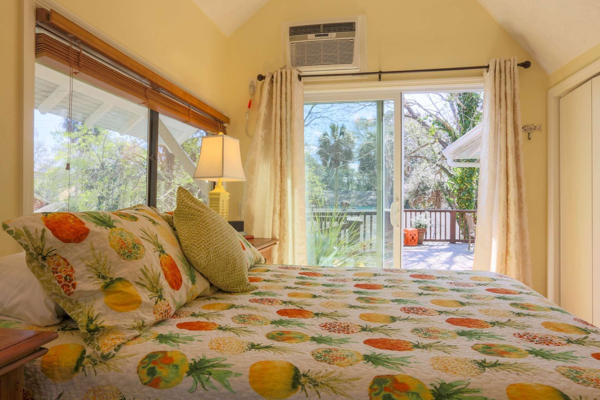 A bedroom with a bed featuring a pineapple-themed bedspread, a window with blinds, a ceiling fan, and a view of a balcony with outdoor furniture.