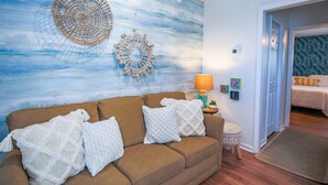 Una sala de estar con un sofá marrón, almohadas blancas de punto, una pared con un mural abstracto azul y blanco, y una mesita redonda con una lámpara.