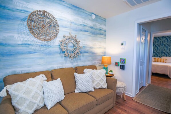 A living room with a brown sofa, white knitted throw pillows, a wall with a blue and white abstract mural, and a small round table with a lamp.
