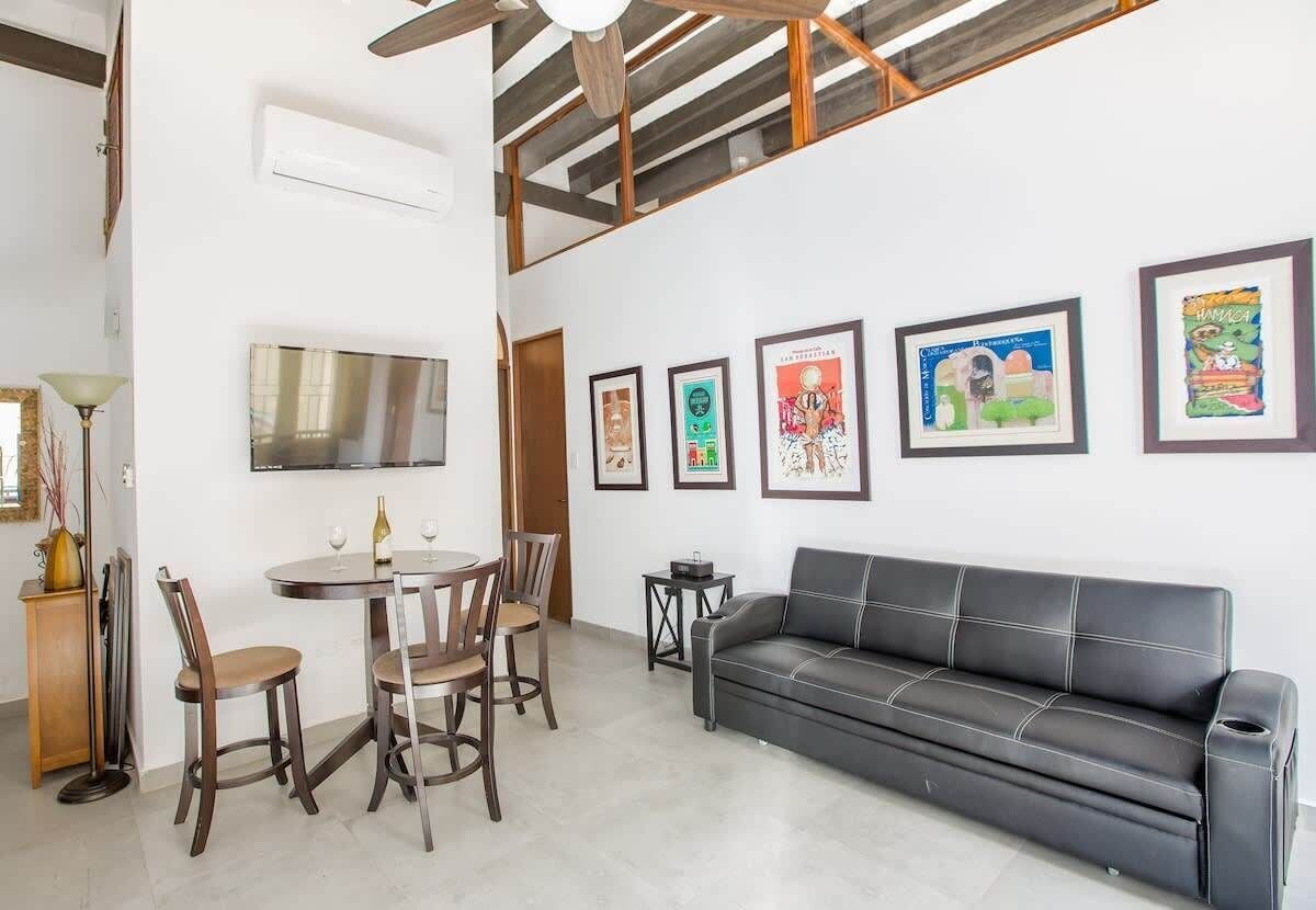 Un salon moderne avec un canapé en cuir noir, une table à manger avec des chaises et un téléviseur à écran plat fixé au mur.