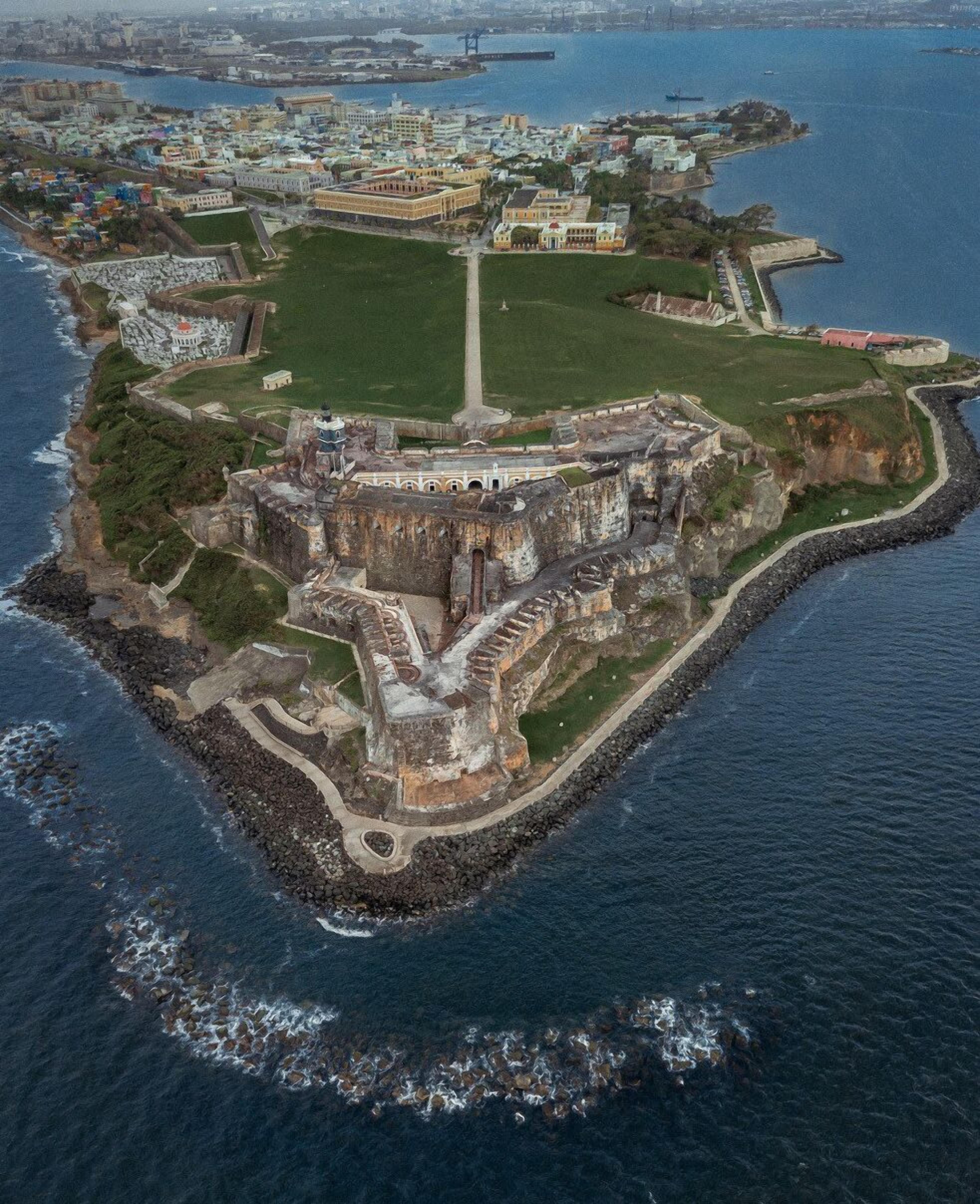 Vista aérea de una fortaleza histórica rodeada por un foso y un paisaje urbano al fondo.