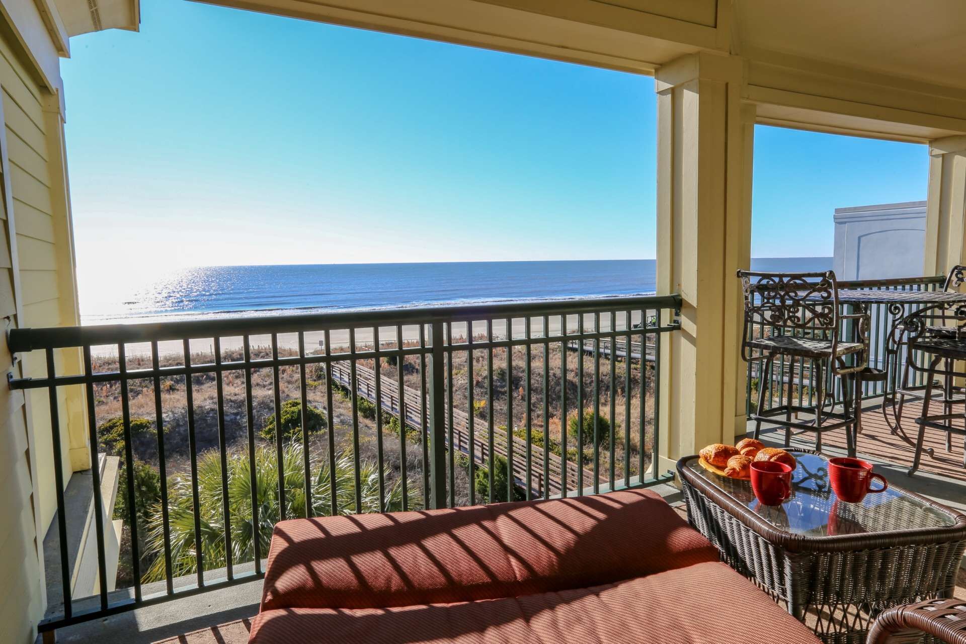 Ein Balkon mit Blick auf den Ozean, ein Korbstuhl mit einem Tablett voller Croissants und ein roter Stuhl.