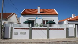 Un bâtiment blanc surmonté d’un toit recouvert de tuiles rouges, une enseigne indiquant « Casa do Bairro by Coca-Cola », et une entrée fermée par un portail, avec deux parasols verts.