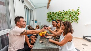 Un groupe de personnes trinquant autour d’une table dont le plateau est en verre, entourée de meubles en osier et d’un mur blanc orné d’une plante verte.