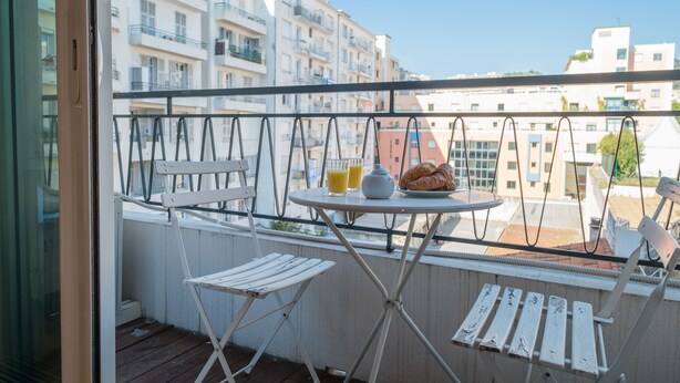 A balcony with a table set for breakfast, including croissants and juice, overlooking a residential area with buildings. - Violet 2 bedroom apartment (Nice)