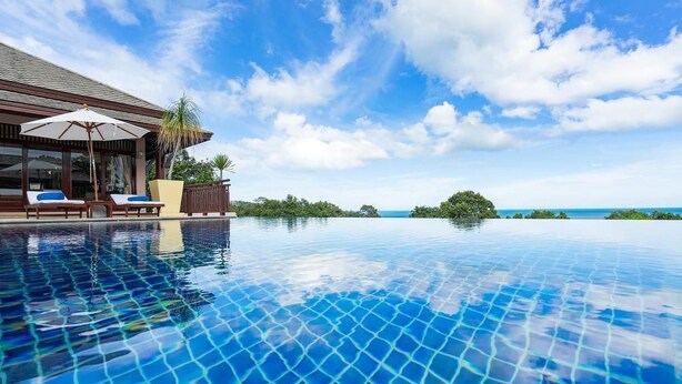 Une piscine à débordement à l’eau d’un bleu cristallin, dotée de chaises longues et de parasols, offrant une vue imprenable sur un paysage tropical et l’océan.