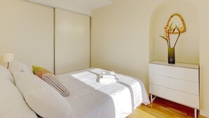 A neatly made bed with white linens and a few pillows, a bedside table with a lamp, and a wall-mounted decorative item. - Yasmin 1 bedroom apartment (Nice)