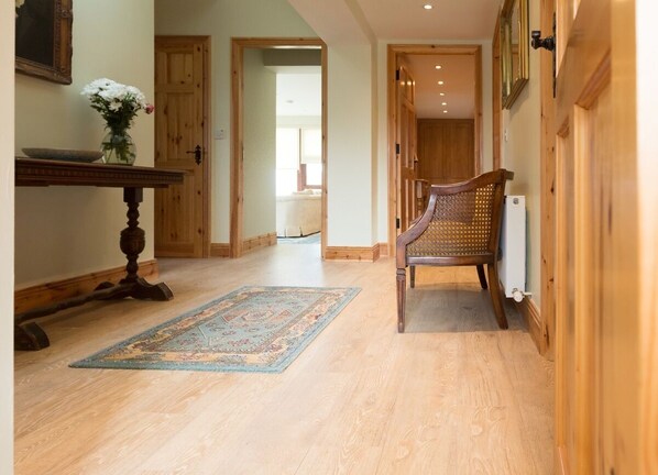 A hallway with wooden flooring, a traditional chair, a table with a vase of flowers, and a rug.
