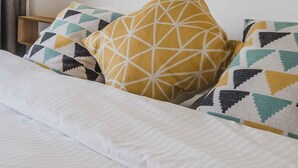 Un llit amb roba de llit blanca, almoinals estampats, un capçal de fusta i una finestra amb cortines.