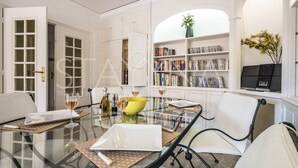Una zona de menjador amb taula de vidre, cadires blanques, un bol groc, copes de vi i una biblioteca amb llibres i un altaveu.