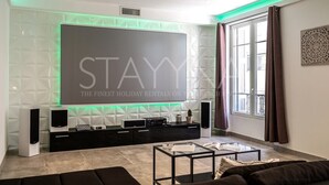 A modern living room with a large flat-screen TV mounted on a geometric-patterned wall, a black entertainment unit, a glass coffee table, and a sofa. - La Carioca (Nice)