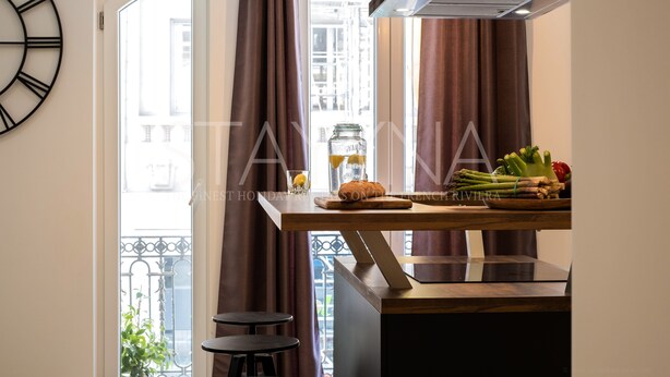A kitchen with a wooden countertop, a stainless steel range hood, a clock on the wall, and a view of the city through large windows. - La Carioca (Nice)