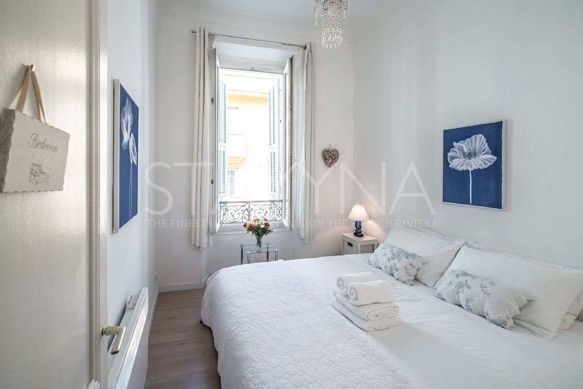 A bedroom with a bed, white bedding, a bedside table with a lamp, a window with curtains, and a wall-mounted heart decoration.