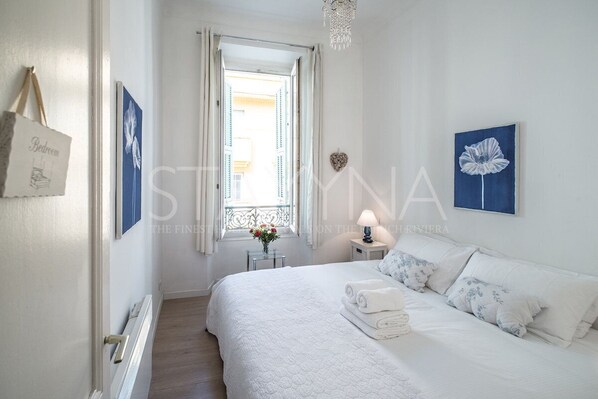 Un dormitorio con cama, ropa de cama blanca, mesita de noche con lámpara, ventana con cortinas y un adorno con forma de corazón en la pared.