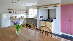 Una cucina con un tavolo da pranzo in legno, sedie e un vaso di fiori. Si nota un fornello, mensole con stoviglie e un armadietto rosa.