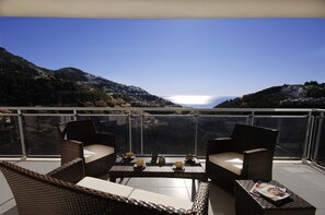 A balcony with wicker furniture, a table with cups and a magazine, and a view of the sea and hills.