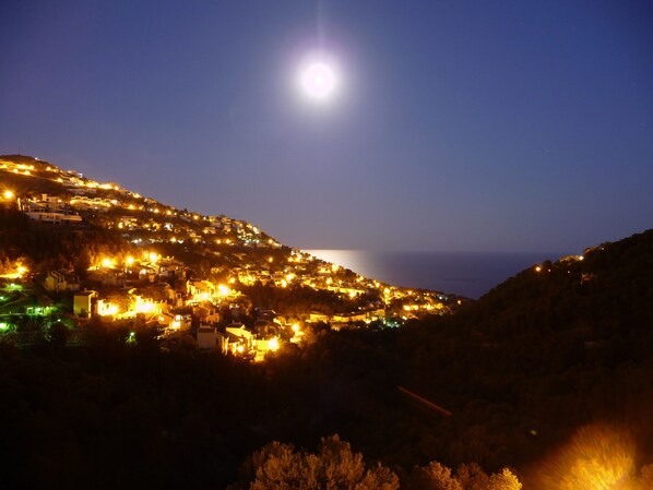 A coastal town at night with illuminated buildings and a visible moon.