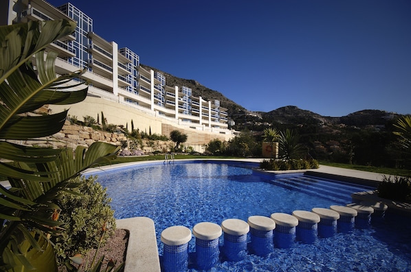 A swimming pool with a clear blue water, surrounded by plants and a building with multiple balconies.