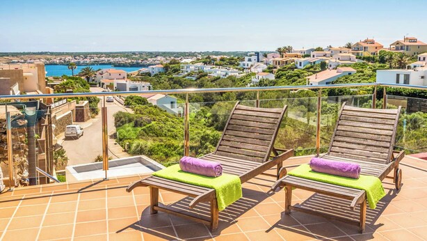 A balcony with two wooden lounge chairs, each with a green towel. The balcony overlooks a coastal town with buildings and greenery.