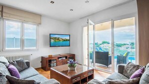 A modern living room with a sofa set, a coffee table, a flat-screen TV, and a sliding glass door leading to a balcony with a view of the sea.